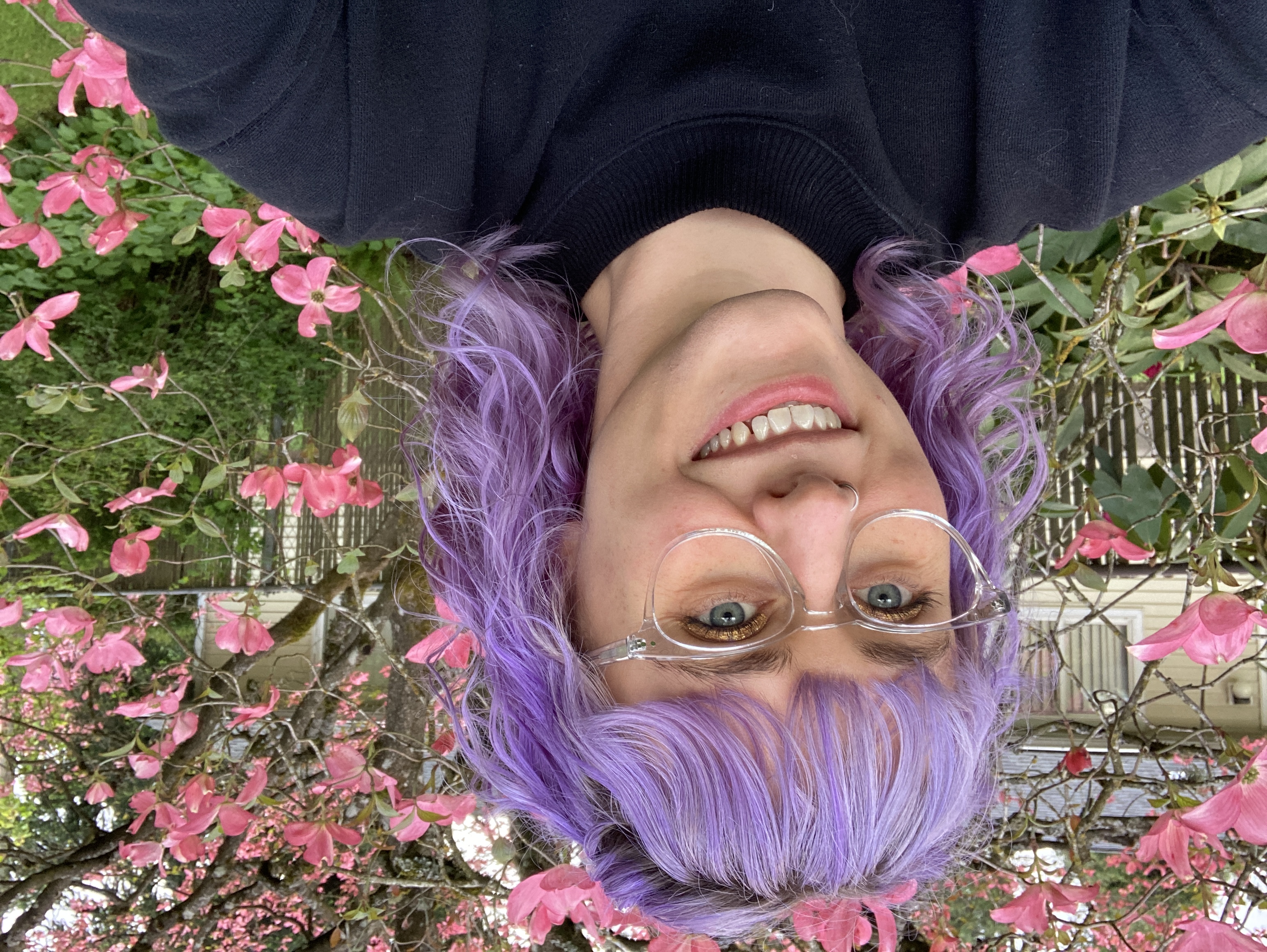 a smiling white person with curly lavendar hair, in the background is pink dogwood trees